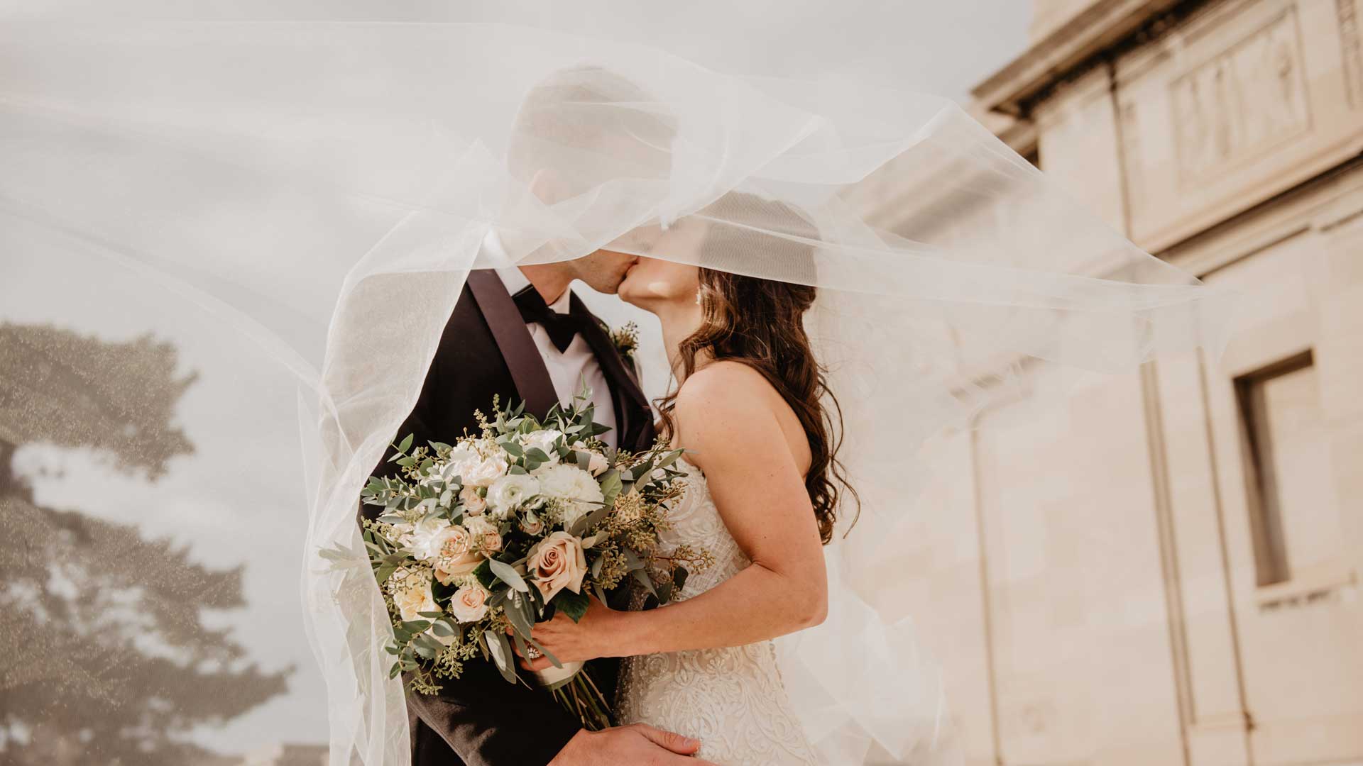 Una novia y un novio se besan bajo un velo delante de un edificio